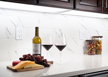 A kitchen counter featuring a white marble backsplash. On the counter are two glasses of wine, an opened bottle of wine, and a cheese and grape charcuterie plate. The photograph is dominantly lit by white LED under-cabinet paneling.
