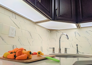 A small assortment of peppers are waiting to be prepped on an off-white marble countertop. The entire kitchen scene is well-lit by LED panel cabinet underlighting.