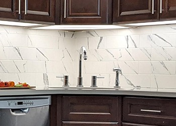 A kitchen sink with chrome hardware. Behind it, the backsplash is white tile with gray marbling. The cabinets are a dark brown wood finish.